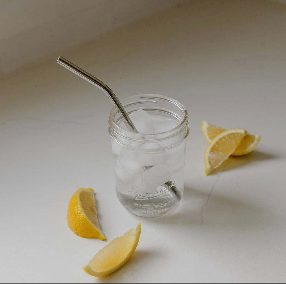 A stainless steel straw sticking out of a glass, filled with ice water, and lemon wedges on the counter next to it.