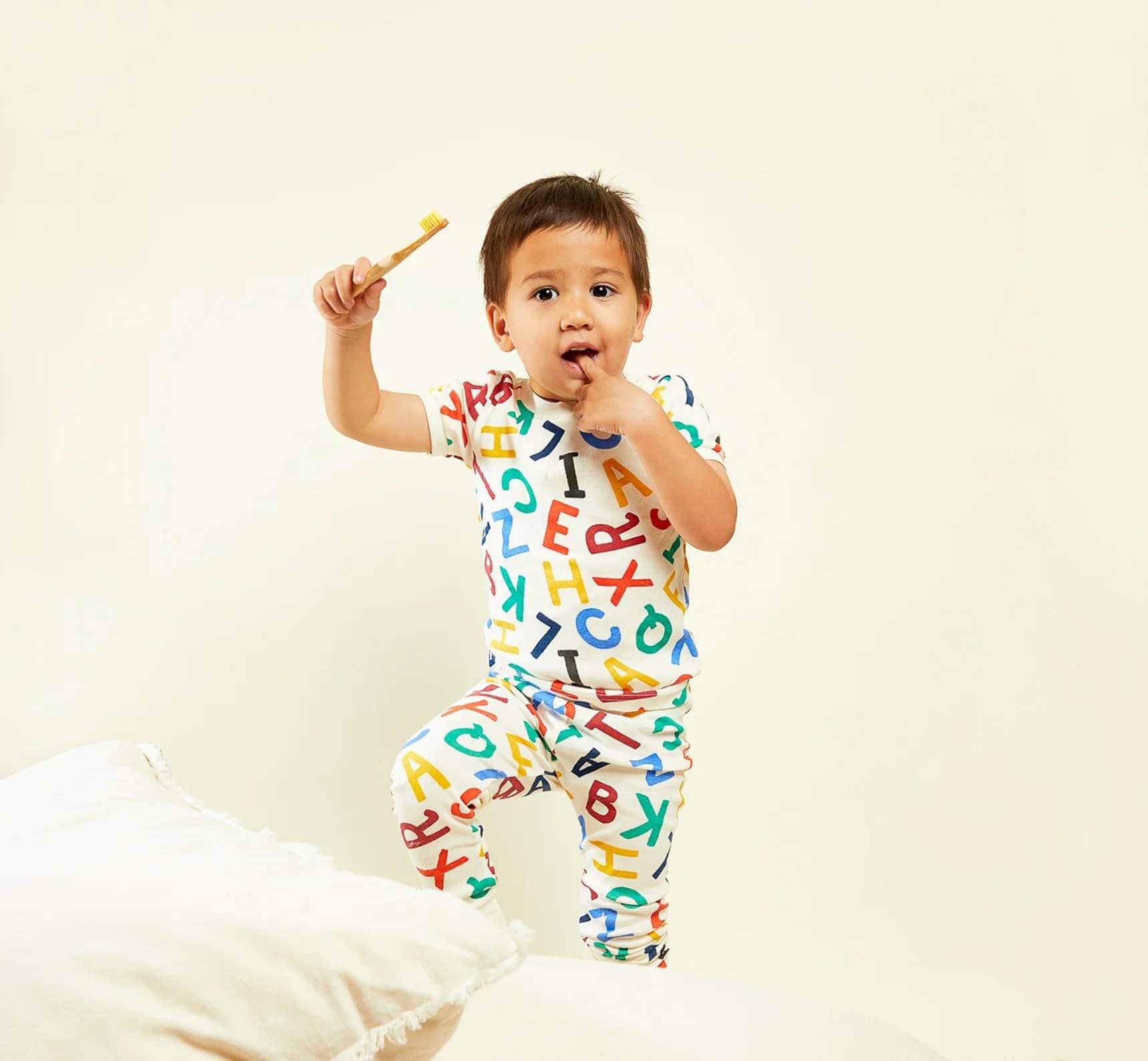 A little boy wearing alphabet pajamas, holding a bamboo toothbrush with his finger in his mouth.