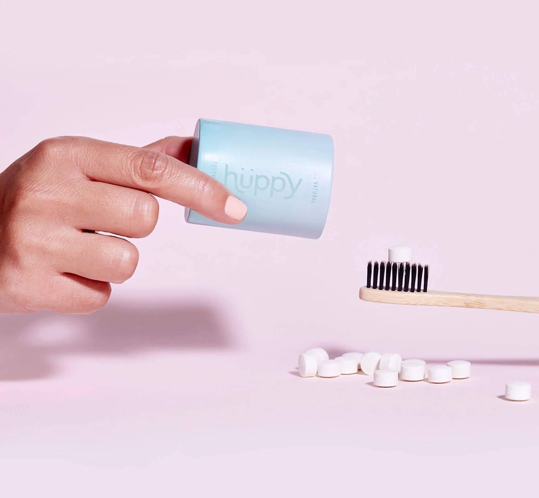 A hand pouring toothpaste tabs out of the blue huppy container and a bamboo toothbrush with a toothpaste tab on it.